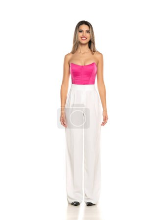 Téléchargez les photos : Jeune femme souriante moderne en pantalon blanc et corset rose posant sur fond de studio blanc - en image libre de droit