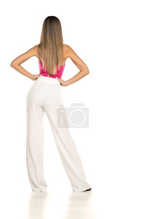 Téléchargez les photos : Vue arrière d'une jeune femme moderne en pantalon blanc et corset rose posant sur fond de studio blanc - en image libre de droit