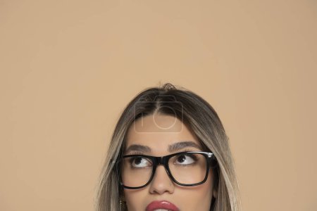 Téléchargez les photos : Demi portrait d'une jeune fille avec des lunettes levant les yeux sur un fond studio beige - en image libre de droit