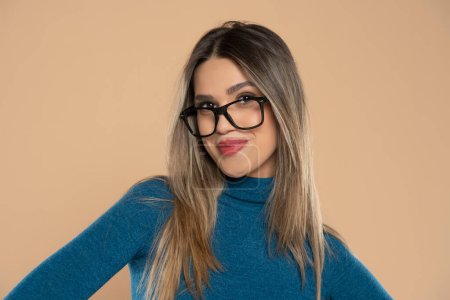 Téléchargez les photos : Jeune femme portant des vêtements décontractés et des lunettes regardant à la caméra, pose de profil relax avec le visage naturel et sourire confiant sur un fond de studio beige - en image libre de droit