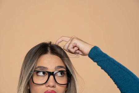 Foto de Medio retrato de una joven con gafas mirando hacia arriba y se rasca la cabeza sobre un fondo de estudio beige - Imagen libre de derechos