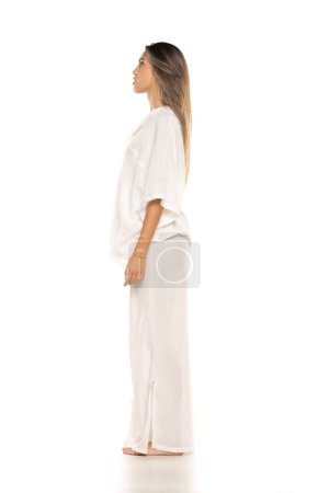 Téléchargez les photos : Jeune femme pieds nus moderne en pantalon blanc et chemisier posant sur fond de studio blanc. vue latérale - en image libre de droit