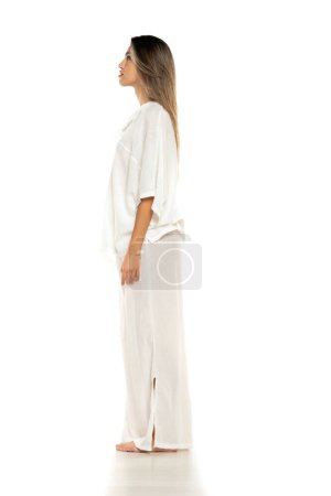 Foto de Mujer descalza moderna joven en pantalones blancos y blusa posando sobre fondo de estudio blanco. vista lateral - Imagen libre de derechos
