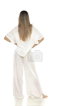 Foto de Mujer descalza moderna joven en pantalones blancos y blusa posando sobre fondo de estudio blanco. atrás, vista trasera - Imagen libre de derechos