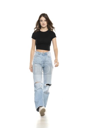 Photo for Young female model wearing ripped jeans and black shirt walking on a white studio background. Front view - Royalty Free Image