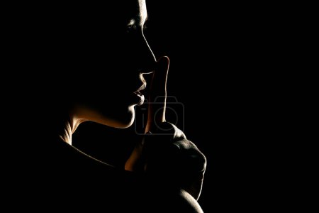 Téléchargez les photos : Silhouette de femme inconnue avec le visage dans l'ombre tenant le doigt sur ses lèvres sur un fond de studio noir - en image libre de droit