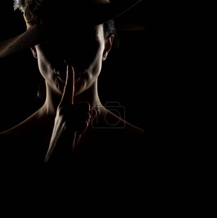 Téléchargez les photos : Silhouette de femme inconnue avec un chapeau, et le visage dans l'ombre tenant doigt sur ses lèvres sur un fond de studio noir - en image libre de droit