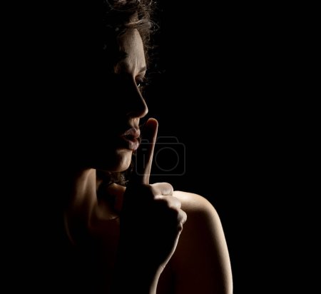 Téléchargez les photos : Silhouette de femme inconnue avec le visage dans l'ombre tenant le doigt sur ses lèvres sur un fond de studio noir - en image libre de droit