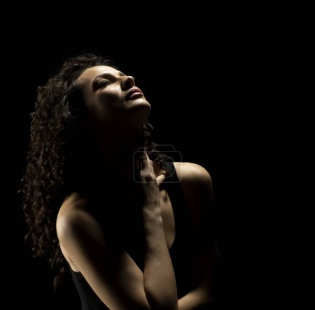 Foto de Retrato de una mujer en la sombra, sujeta su cuello, ojos cerrados sobre un fondo negro del estudio - Imagen libre de derechos