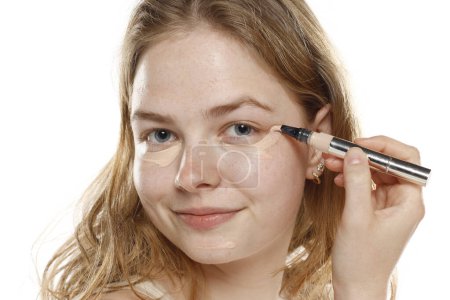 Foto de Mujer rubia joven aplicar corrector bajo el ojo en un fondo de estudio blanco - Imagen libre de derechos