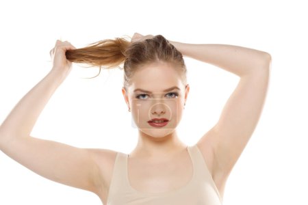 Téléchargez les photos : Vue de face d'une jeune femme blonde attachant ses longs cheveux sur un fond de studio blanc - en image libre de droit