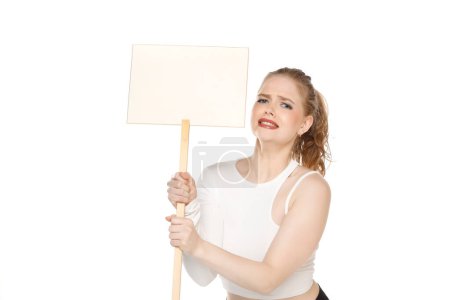 Téléchargez les photos : Jeune femme blonde malheureuse tenant signe blanc sur fond de studio blanc. - en image libre de droit