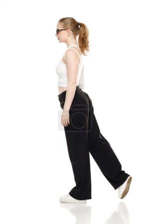 Photo for Side view of young blond woman in trousers and white blouse walking on white studio background - Royalty Free Image