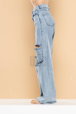 Téléchargez les photos : Jambes d'une femme en jean déchiré sur fond studio beige. Côté, vue de profil. - en image libre de droit