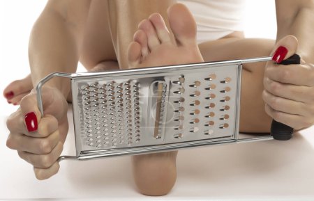 Téléchargez les photos : Une femme se gratte les pieds avec une râpe de cuisine sur fond de studio blanc - en image libre de droit