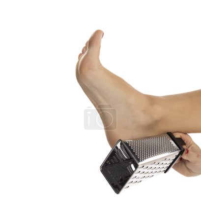 Téléchargez les photos : Une femme se gratte les pieds avec une râpe de cuisine sur fond de studio blanc - en image libre de droit