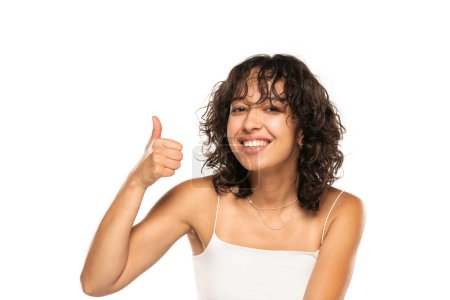 Téléchargez les photos : Jeune femme souriante à la peau foncée sans maquillage posant sur un fond de studio blanc. Pouce en l'air. - en image libre de droit