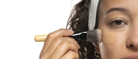 Téléchargez les photos : Jeune femme etnic applaudit fonderie liquide sur son visage avec pinceau de maquillage sur fond de studio blanc - en image libre de droit