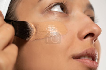 Téléchargez les photos : Jeune femme etnic applaudit fonderie liquide sur son visage avec pinceau de maquillage sur fond de studio blanc - en image libre de droit