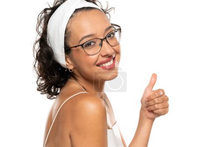 Foto de Joven mujer sonriente de piel oscura con maquillaje, diadema y gafas posando sobre un fondo de estudio blanco. Pulgares arriba. - Imagen libre de derechos