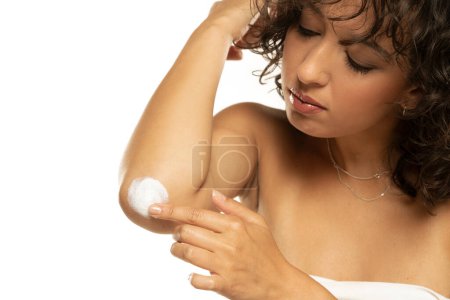 Téléchargez les photos : Prise de vue de tête d'une femme exotique prend soin de ses coudes à l'aide de crème cosmétique sur un fond de studio blanc - en image libre de droit