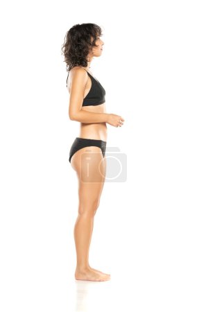 Foto de Joven mujer morena exótica en bikini negro posando sobre un fondo de estudio blanco. Perfil, vista lateral. - Imagen libre de derechos