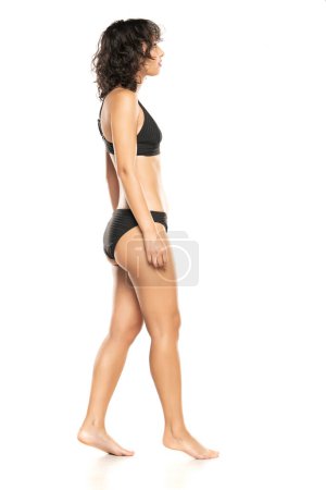 Foto de Joven mujer morena exótica en traje de baño bikini negro caminando sobre un fondo de estudio blanco. Perfil, vista lateral. - Imagen libre de derechos