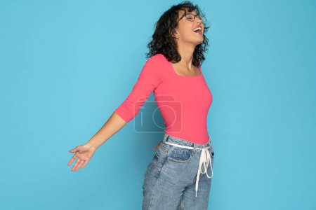 Téléchargez les photos : Jeune femme brune exotique heureuse en chemisier rose et jean ceinture de corde posant sur un fond de studio blanc. Vue latérale. - en image libre de droit