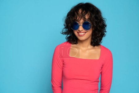 Photo for Young exotic smiling brunette woman in pink blouse posing with a sunglasses on a blue studio background - Royalty Free Image