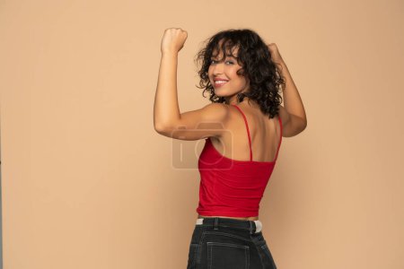 Téléchargez les photos : Jeune femme brune exotique heureuse en chemise et jeans sans manches rouges montrant des biceps sur un fond studio beige - en image libre de droit
