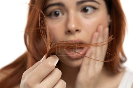 Foto de Mujer pelirroja preocupada inspeccionando sus puntas de pelo en un fondo de estudio blanco - Imagen libre de derechos