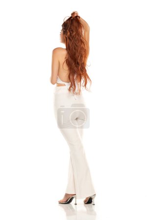 Photo for Fashion style rear view studio shott of beautiful young redhead woman. White wide leg pants and white sleevles shirt. Model standing and posing against white studio background - Royalty Free Image