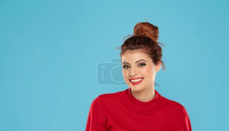 Photo for Cheerful gorgeous young woman wearing her ginger hair in bun smiling happily dressed in red blouse looking at camera with excited joyful smile on a blue studio background - Royalty Free Image