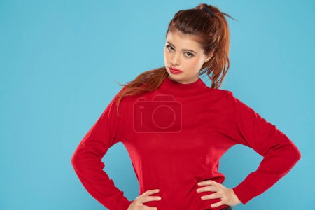 Téléchargez les photos : Malheureuse jeune femme portant ses cheveux roux en queue de cheval vêtue d'un chemisier rouge regardant la tristesse de la caméra sur un fond bleu studio - en image libre de droit