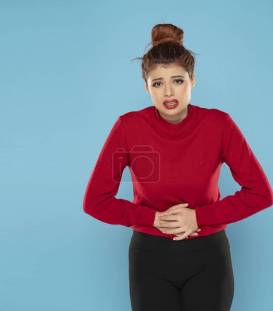 Téléchargez les photos : Femme rousse souffrant de douleurs à l'estomac et tenant ses mains dans le bas-ventre sur un fond de studio bleu - en image libre de droit