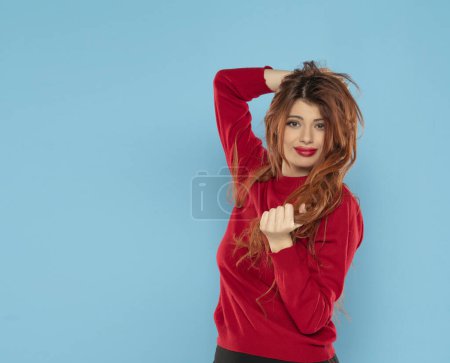 Téléchargez les photos : Jeune femme confus avec de longs cheveux gingembre désordonné habillé en chemisier rouge en regardant la caméra sur un fond de studio bleu - en image libre de droit