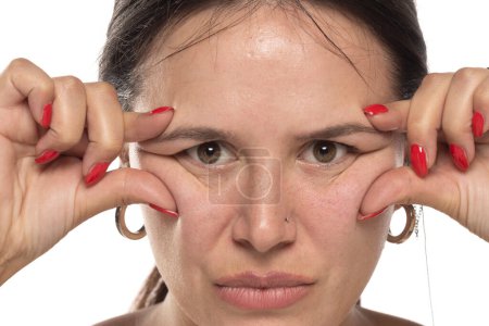 Téléchargez les photos : Femme pincant les rides de ses yeux avec ses doigts sur un fond de studio blanc - en image libre de droit