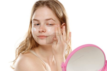 Téléchargez les photos : Souriante jeune femme blonde appliquant une crème sur son visage sur un fond de studio blanc - en image libre de droit