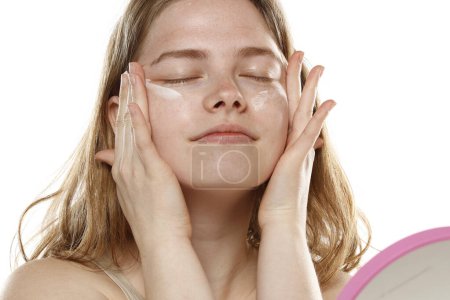 Téléchargez les photos : Jeune femme blonde positive appliquant une crème sur son visage sur un fond de studio blanc - en image libre de droit