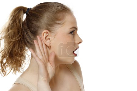Téléchargez les photos : Jeune femme blonde caucasienne écoutant quelque chose en mettant la main sur l'oreille sur un fond de studio blanc. Vue du profil - en image libre de droit