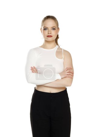 Téléchargez les photos : Vue de face de la jeune femme blonde en pantalon et blouses blanches posant les bras croisés sur fond de studio blanc - en image libre de droit