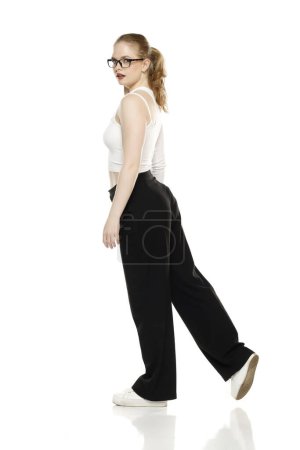 Foto de Vista lateral de la joven rubia en pantalones y blusa blanca caminando sobre fondo blanco del estudio - Imagen libre de derechos