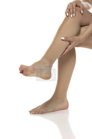 Téléchargez les photos : Une femme appliquant de la lotion et massant sa jambe sur fond de studio blanc, favorisant l'autosoin et la détente. Concept d'autosoin et routine de soins de la peau. - en image libre de droit