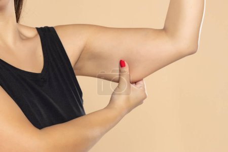 Foto de Primer plano de una mujer agarrando la piel en la parte superior del brazo con exceso de grasa aislada en un fondo de estudio beige. Pellizcar los músculos sueltos y caídos. Concepto de sobrepeso - Imagen libre de derechos
