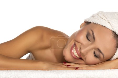 Téléchargez les photos : Portrait de beauté d'une jolie femme à moitié nue gaie avec une serviette enroulée autour de sa tête avec le sourire des dents et les yeux fermés couchés sur fond de studio blanc - en image libre de droit