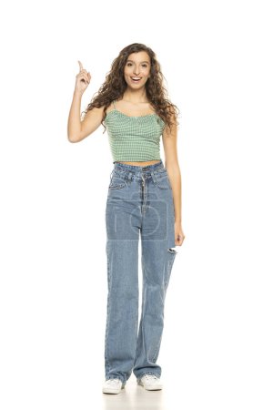 Photo for Full length shot of young smiling woman wearing green shirt and blue jeansi while standing at isolated white studio background and pointing up. - Royalty Free Image