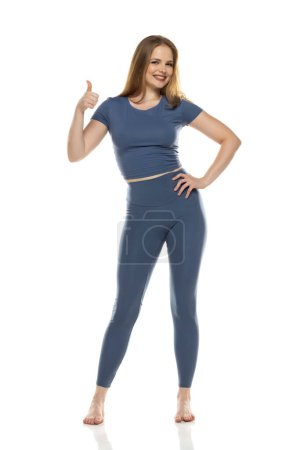 Photo for Full body, front view young smiling woman in sportswear posing on white studio background and showing thumbs up - Royalty Free Image