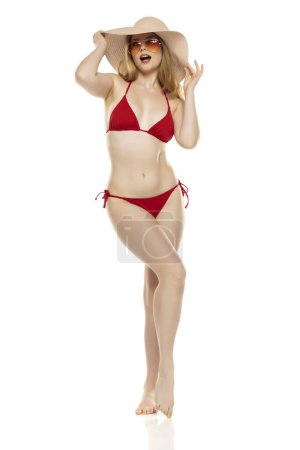 Foto de Cuerpo completo, vista frontal joven rubia en bikini, gafas de sol y sombrero posando sobre fondo blanco del estudio - Imagen libre de derechos