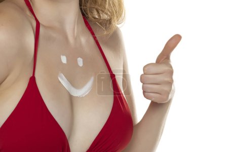 Foto de Pecho femenino en sujetador rojo con producto cosmético en forma de sonrisa sobre fondo blanco del estudio.Pulgares hacia arriba! - Imagen libre de derechos