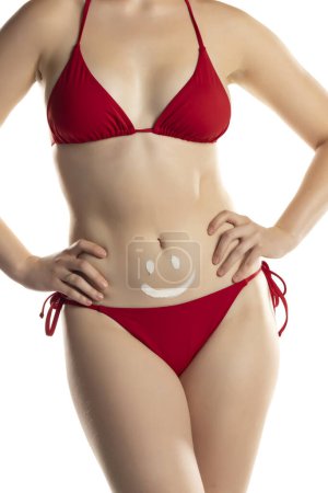 Photo for Young woman in red bikini with cosmetic product in smile shape on her belly on white studio background. - Royalty Free Image